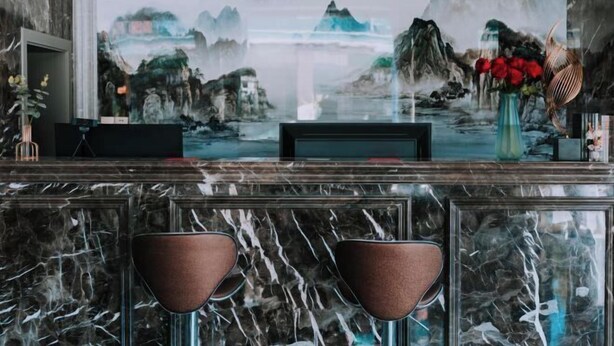 Un comptoir d’accueil avec un dessus en marbre, deux fauteuils marron à coussins, un vase rempli de fleurs rouges et un grand tableau représentant un paysage montagneux.