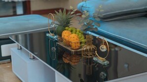 Une chambre moderne avec une table à plateau en verre sur laquelle se trouvent un panier de fruits, un ananas et une plante décorative.