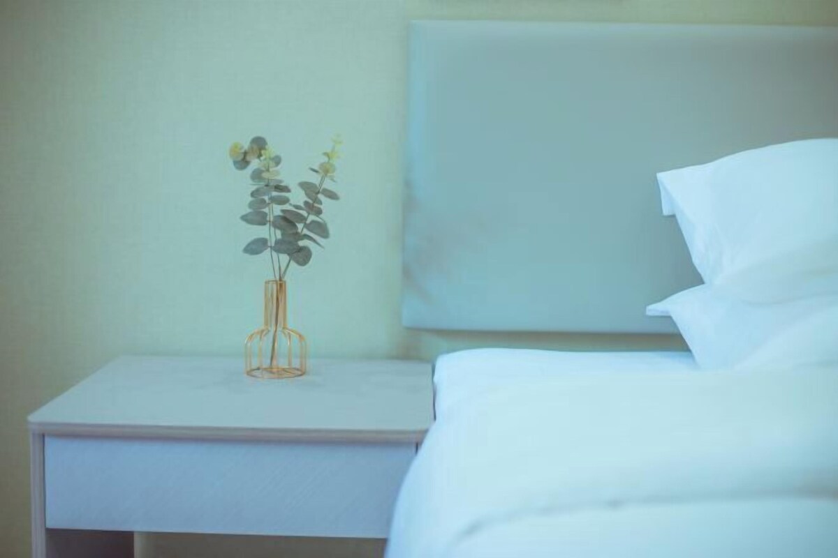 Una cama bien hecha con sábanas blancas, una mesita de noche blanca con un florero de vidrio transparente que contiene una planta, y una pared lisa de fondo.