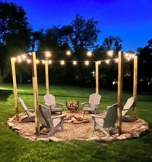 Un gazebo con luci a stringa, circondato da poltrone Adirondack e un braciere, immerso in un'area erbosa.