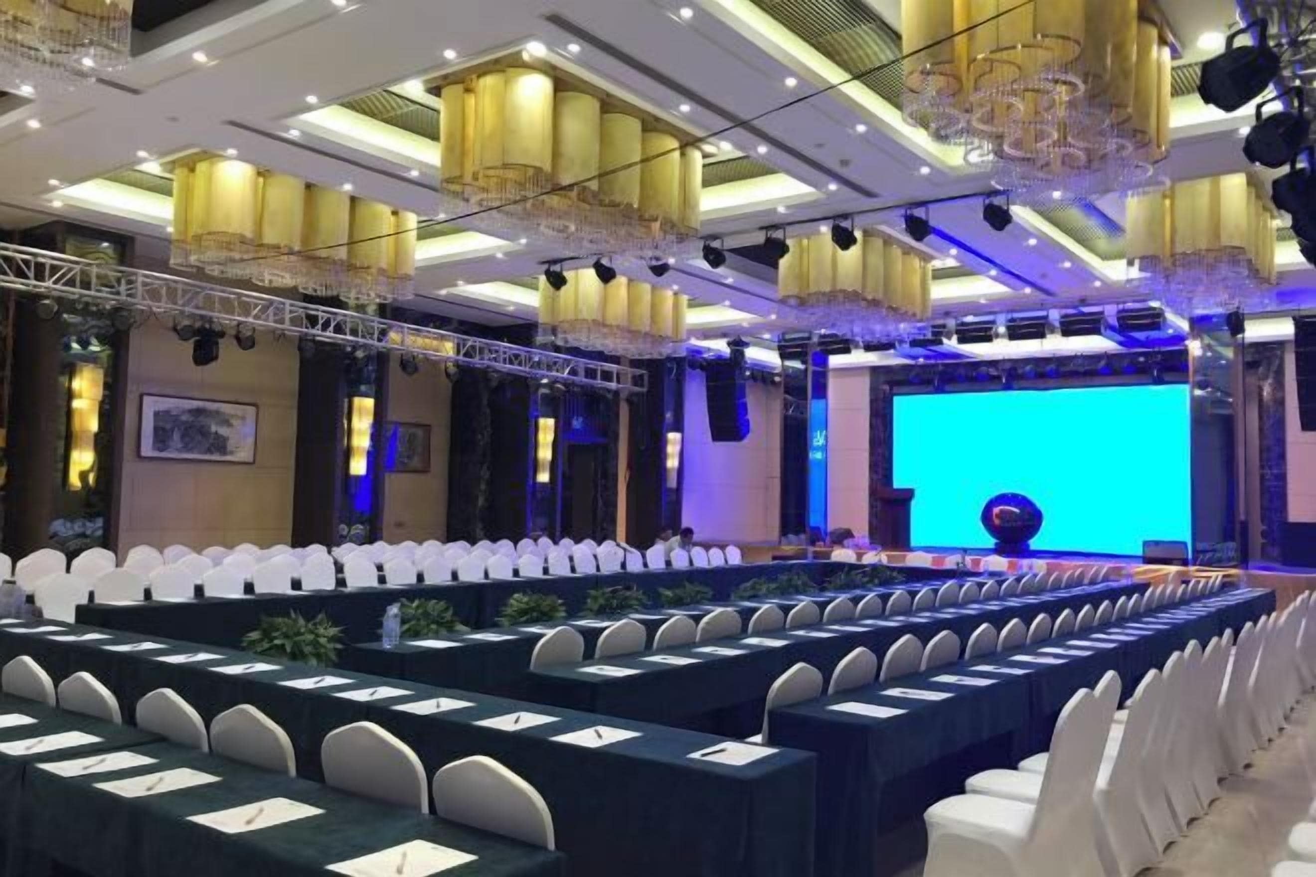 A conference room with rows of tables and chairs, a large screen, and stage lighting.