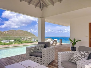 A covered outdoor area with a pool, two wicker chairs with cushions, a potted plant, and a view of the sea and hills.