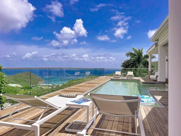 A wooden deck with a pool, lounge chairs, and a view of the ocean.