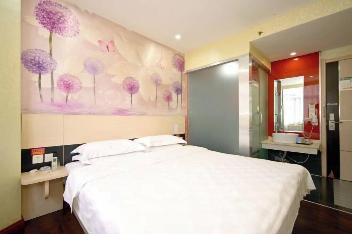 A hotel room with a bed, a wall-mounted painting, a bedside table, a mirror, and a bathroom visible through an open door.