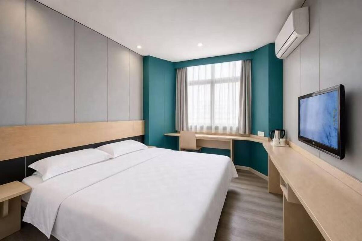 A hotel room with a bed, a desk, a chair, a television, and an air conditioning unit.