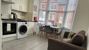 Un espace de vie compact comprenant un coin cuisine, un lave-linge, un coin repas avec des chaises et un canapé avec une télécommande posée sur une petite table.