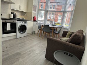 A compact living space with a kitchenette, a washing machine, a dining area with chairs, and a sofa with a remote control on a small table.