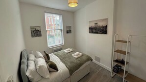 Une chambre à coucher avec un lit, une fenêtre avec des stores, une étagère fixée au mur et un luminaire au plafond.