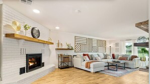 A living room with a white brick fireplace, a sectional sofa, and a wooden floor. - Sleeps 22—Pickleball~Hot Tub~Pool Table~Games (Broken Arrow)