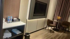 Ein Hotelzimmer mit Flachbildfernseher, zwei Sesseln, einem kleinen Tisch mit einer Vase, einer Ablage mit Handtüchern und einem Mülleimer.
