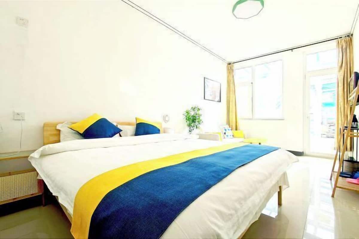 Une chambre d’hôtel avec un lit, une table de chevet, une lampe, une fenêtre avec des rideaux et une plante.