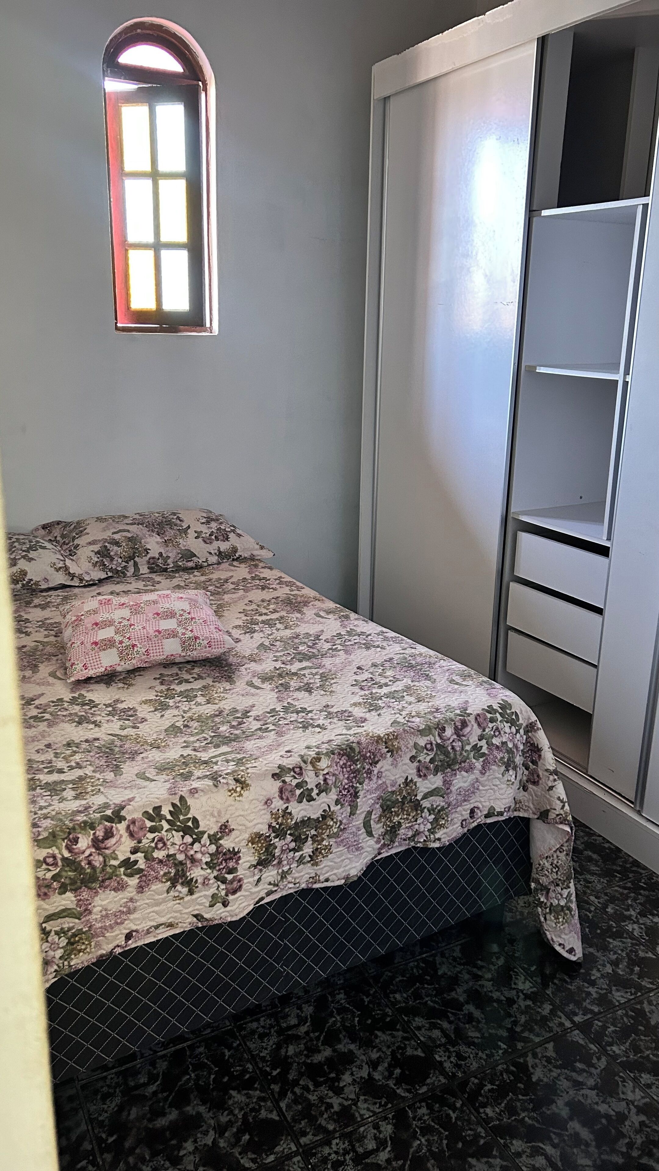 Un dormitorio con cama, armario, ventana pequeña y suelo de baldosas.