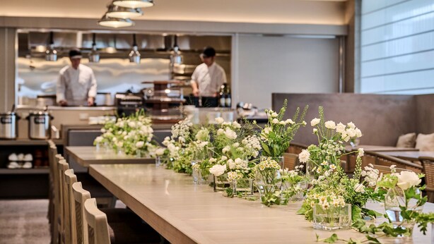 Ein langer Esstisch mit floralen Mittelpunkten, ein Küchenbereich mit Köchen und ein modernes Interieur.