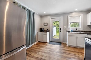 A modern kitchen with a stainless steel refrigerator, white cabinets, a window with blinds, and a door leading outside.