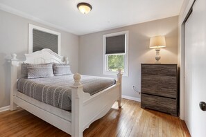 A bedroom with a bed, two windows with blinds, a dresser, and a lamp.
