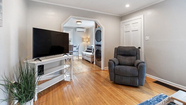 Una sala de estar con un televisor de pantalla plana sobre un mueble blanco, un sillón gris y piso de madera.