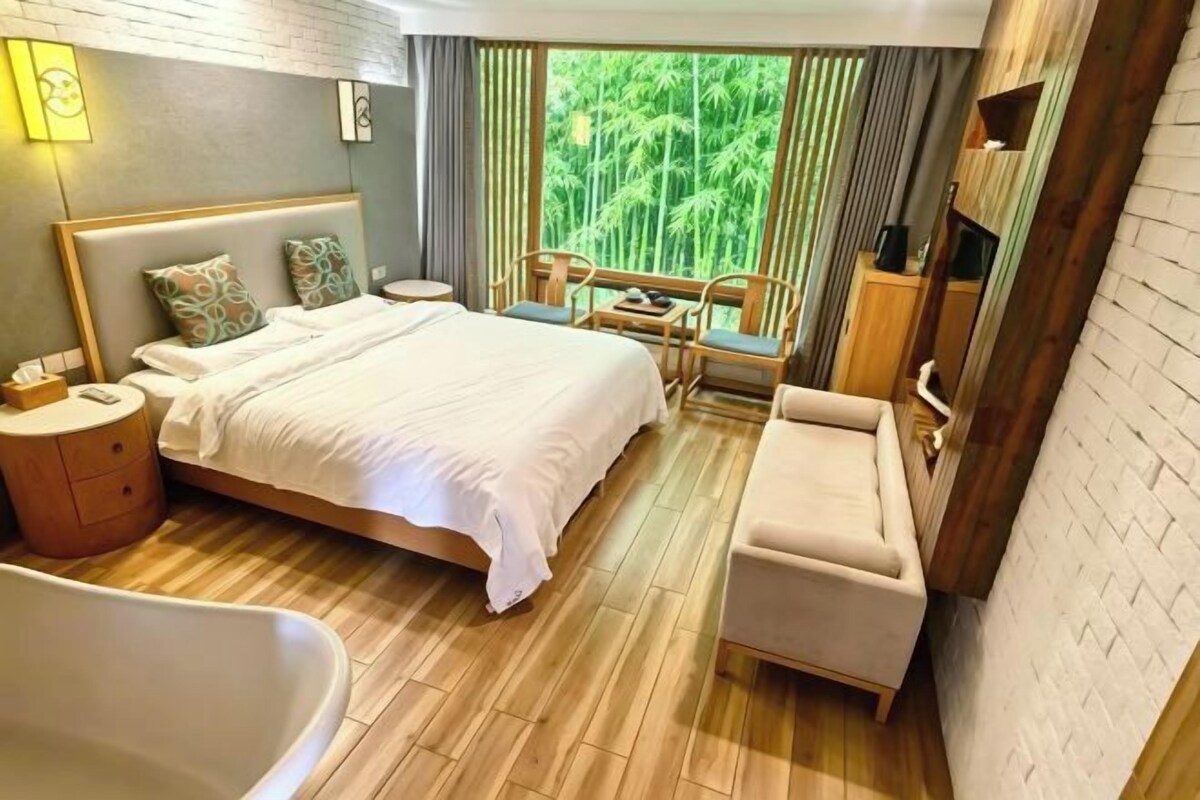 A hotel room with a large bed, a sitting area with a table and chairs, a bathtub, and a view of greenery outside.