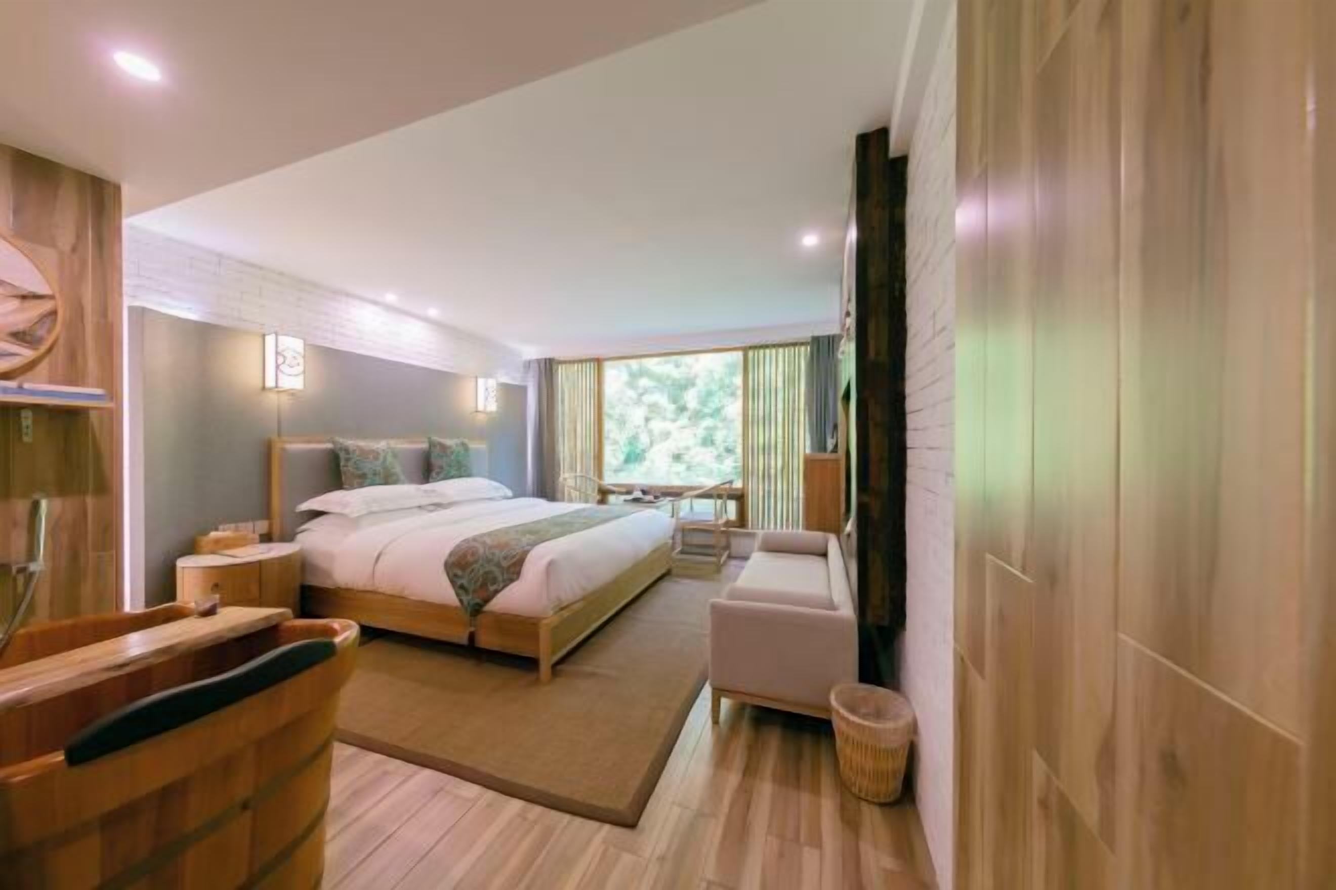 Habitación de hotel moderna con una cama grande, una zona de estar, una bañera de madera y vistas a zonas verdes al exterior.