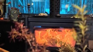 A wood-burning stove with a visible fire, surrounded by decorative items and a window with a blue curtain. - Wuxi Dayin Mountain Residence Garden Homestay (Wuxi)
