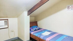 Une petite chambre avec un lit simple, une tête de lit en bois, un lit recouvert d’une couverture à rayures bleues et roses, et un panneau « Il est interdit de fumer » accroché au mur.