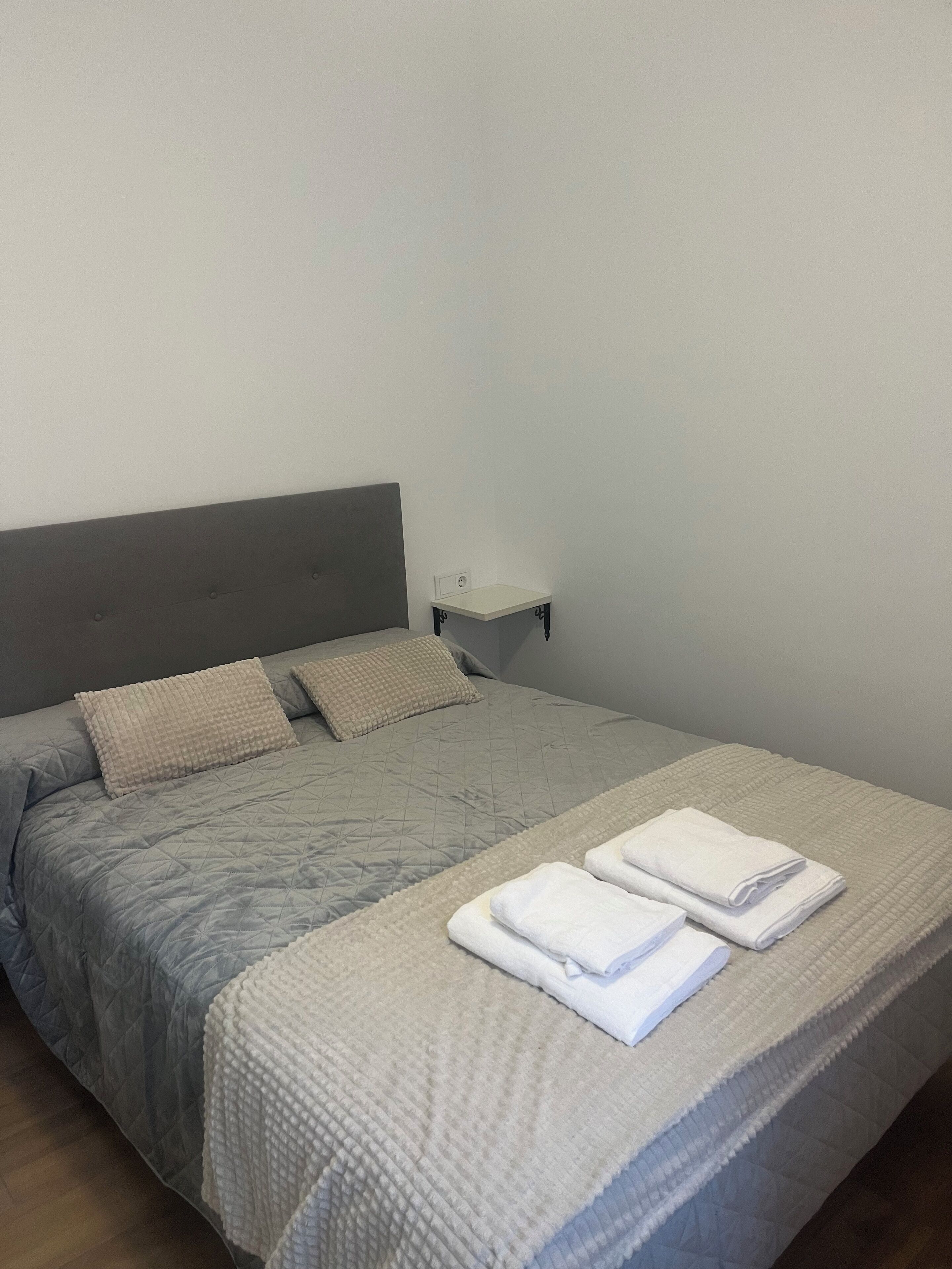 A neatly made bed with a grey quilt, two pillows, and folded white towels on the bed.