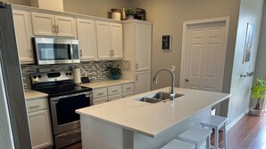 A kitchen with white cabinets, a stainless steel refrigerator, a built-in oven, a microwave, and a sink. There are two white barstools and a potted plant. - 4 Bedroom with all the things! (Eugene)