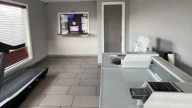 A modern, well-lit room with a treadmill, a large window with blinds, a flat-screen TV mounted on the wall, and a kitchenette area. - Westerner Motel (Port Allen)