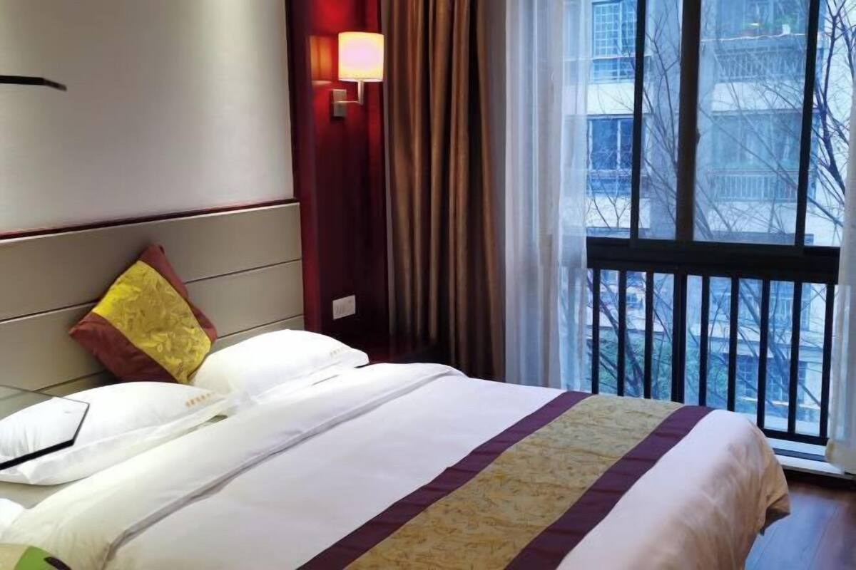 A hotel room with a bed, a nightstand, a lamp, a window with a view of buildings, and a balcony railing.