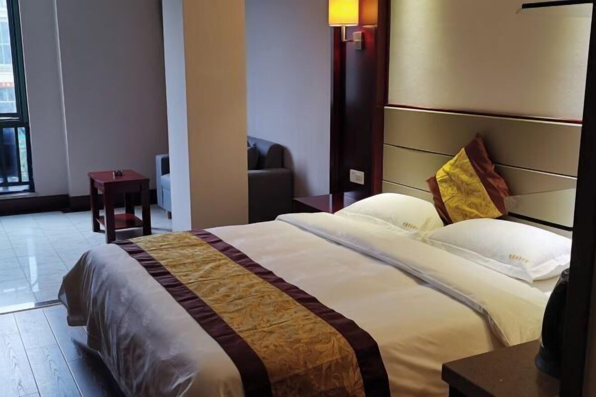 A hotel room with a large bed, a small table, a chair, and a wall-mounted lamp.