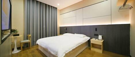 Una habitación de hotel moderna con una cama grande, mesitas de noche, una pequeña zona de estar y un ventanal con cortinas.