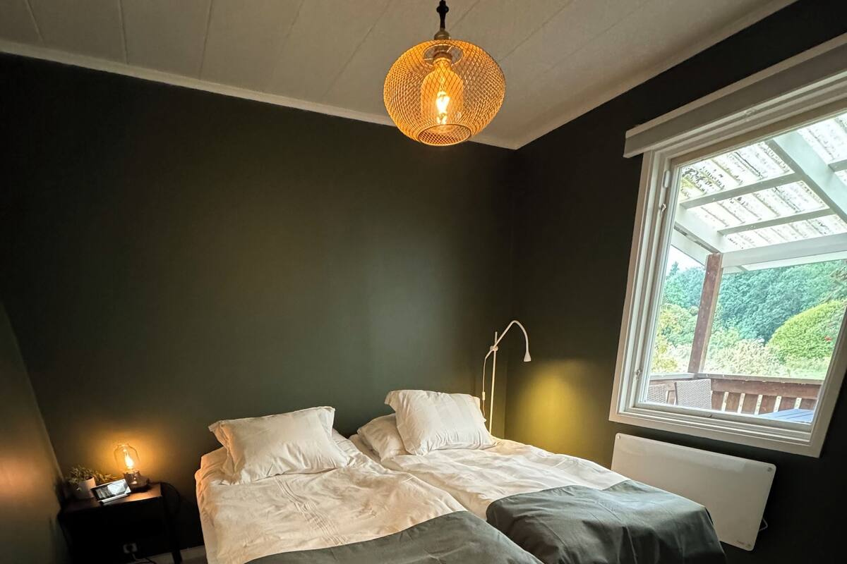 A bedroom with two beds, a nightstand, a lamp, and a window with a view of greenery.