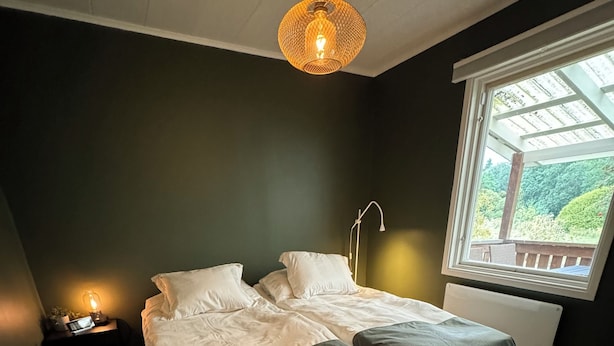 A bedroom with two beds, a nightstand, a lamp, and a window with a view of greenery.