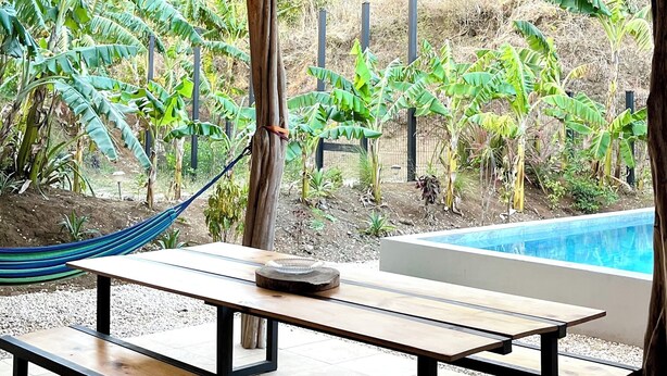 An outdoor patio with a wooden table and benches, a hammock, and a view of a swimming pool and greenery. - CASA ULYSSE 
 2-bedroom house near Tamarindo with swimming pool in the nature (Tamarindo)