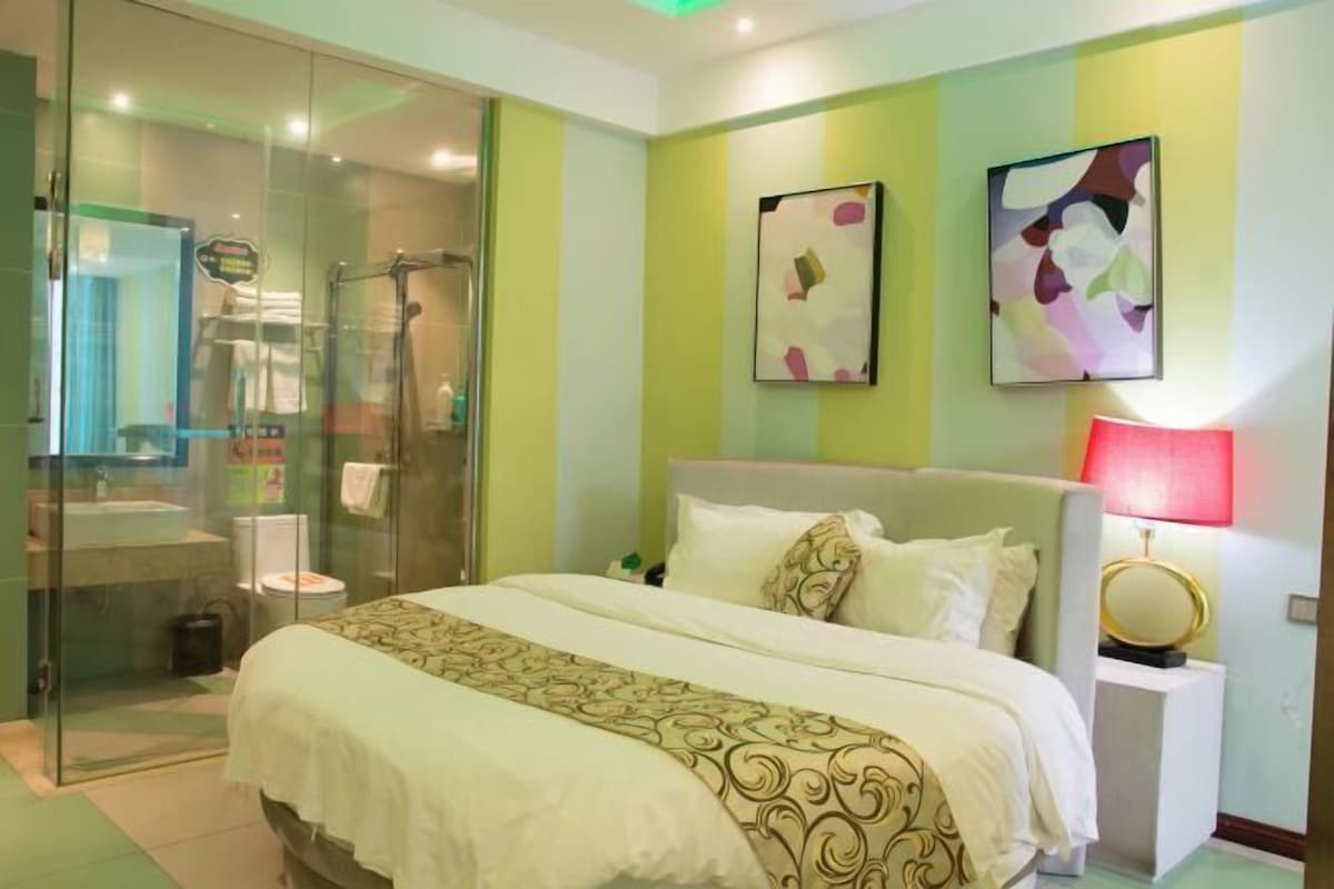 A hotel room with a bed, a bedside table, a nightstand, a glass shower enclosure, and a bathroom with a toilet and sink.