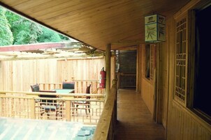 Terraza de madera con mesa y sillas, un extintor y un corredor cubierto que conduce a un edificio.