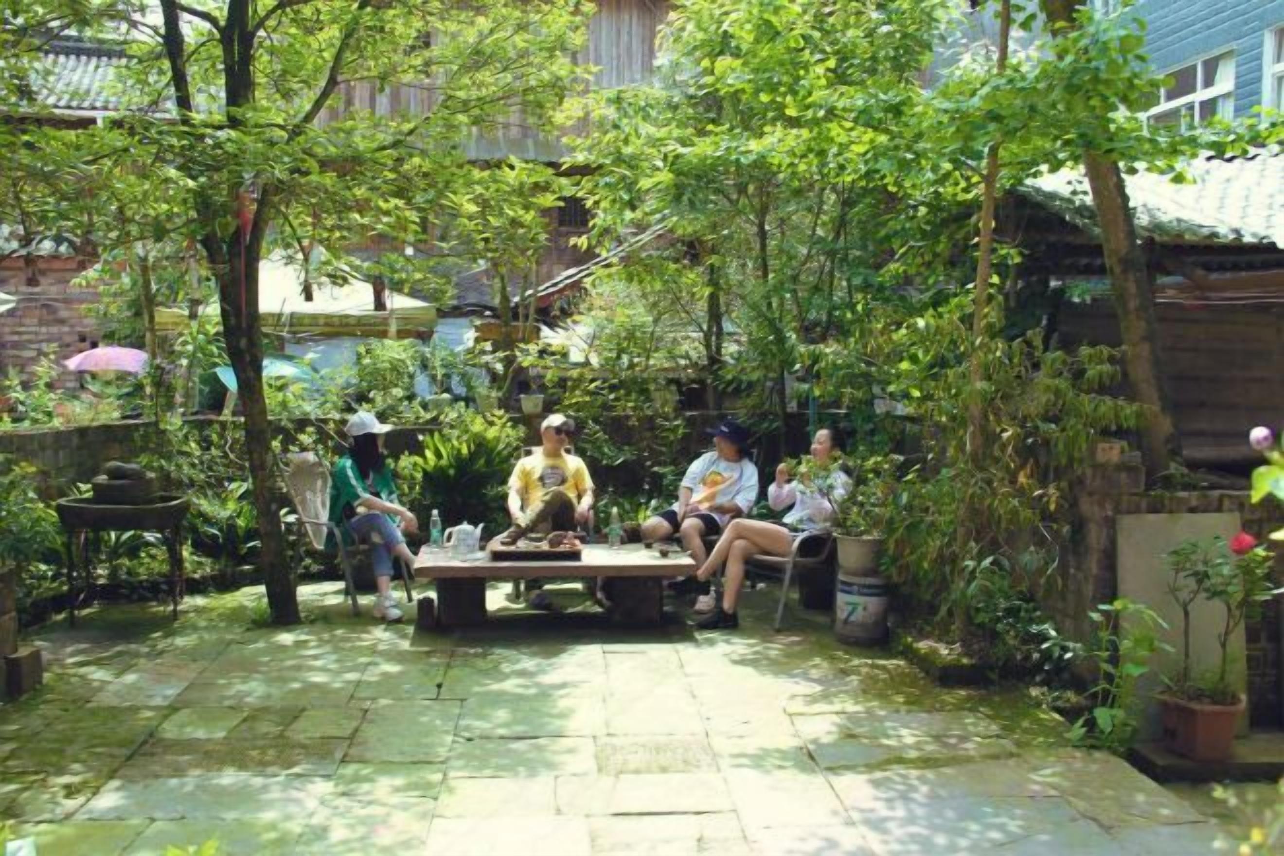 Un grup de persones assegudes al voltant d'una taula de fusta en un jardí amb plantes en testos i un mur de maó al fons.