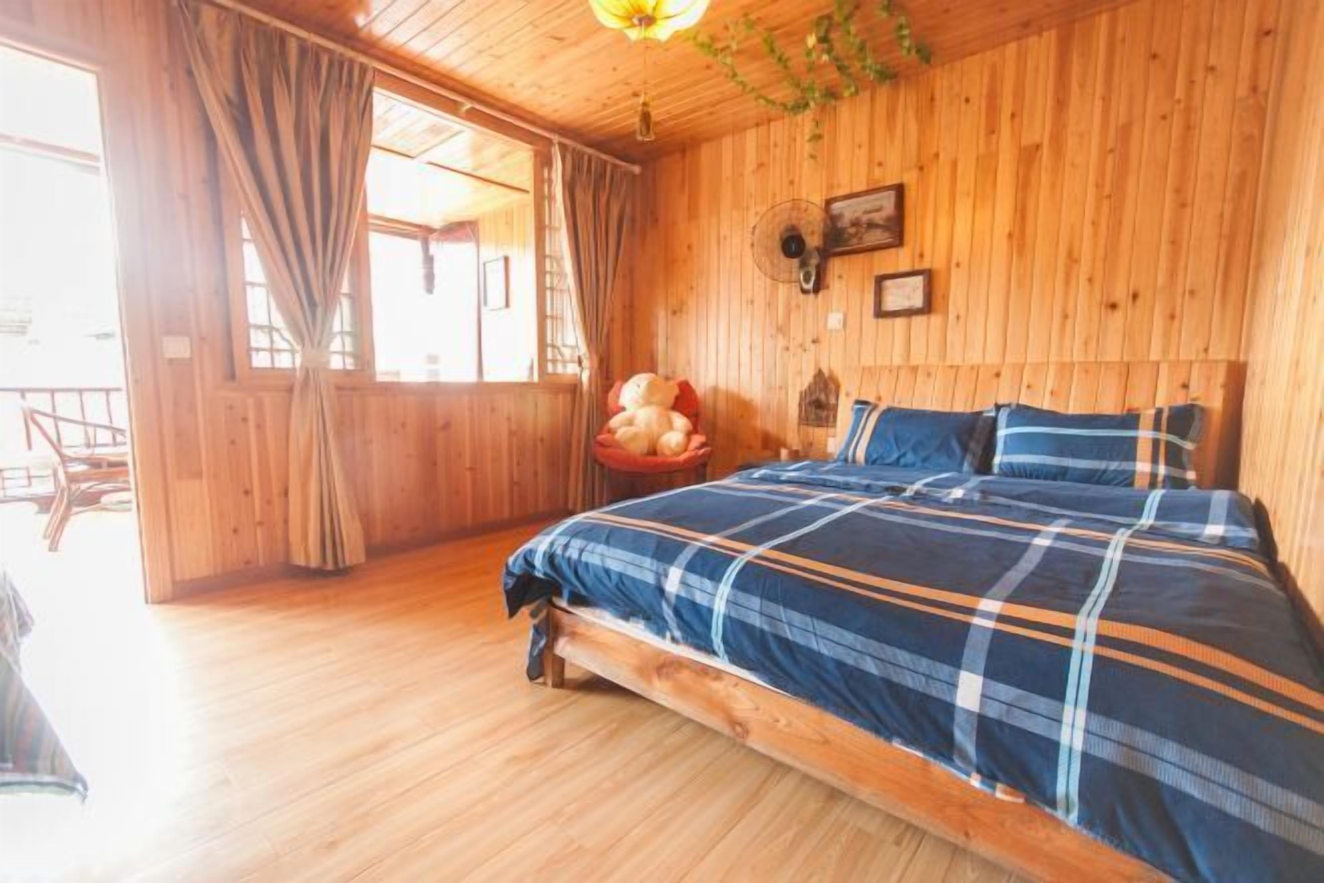 A wooden room with a bed, a window with curtains, and a fan on the wall.