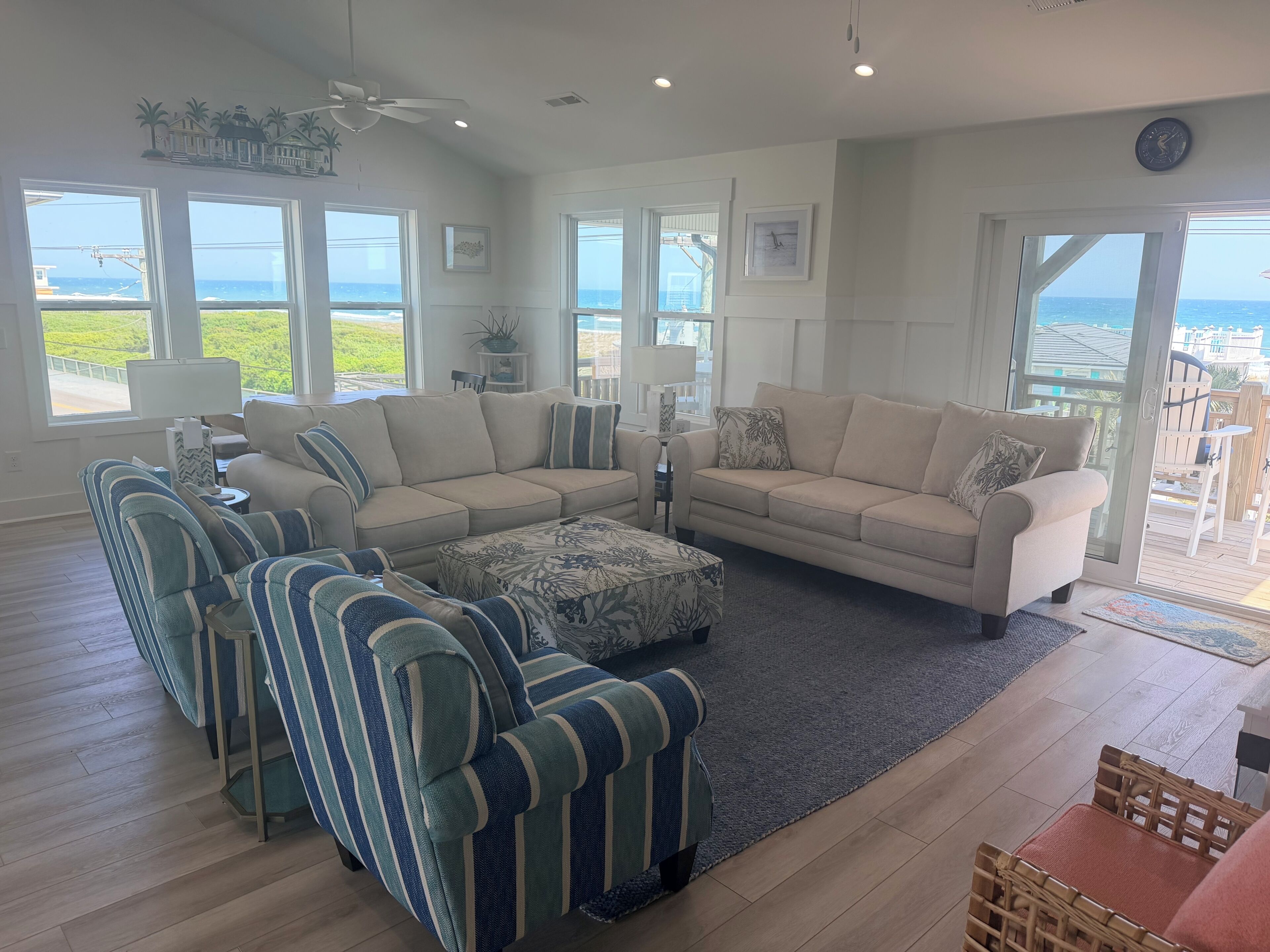 Ein geräumiges Wohnzimmer mit großem Fenster und Meerblick, einem Deckenventilator sowie Blick auf das Meer durch die Glastür.