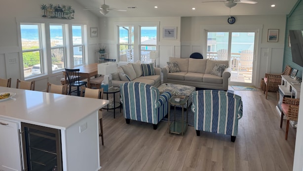 A spacious living room with a large window overlooking the beach, a cozy seating area with striped ottomans, and a dining area with a wooden table and chairs. - Bright Beach Escape • Ocean Views • Easy Beach Access (Topsail Beach,)