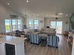 A spacious living room with a large window overlooking the beach, a cozy seating area with striped ottomans, and a dining area with a wooden table and chairs. - Bright Beach Escape • Ocean Views • Easy Beach Access (Topsail Beach,)