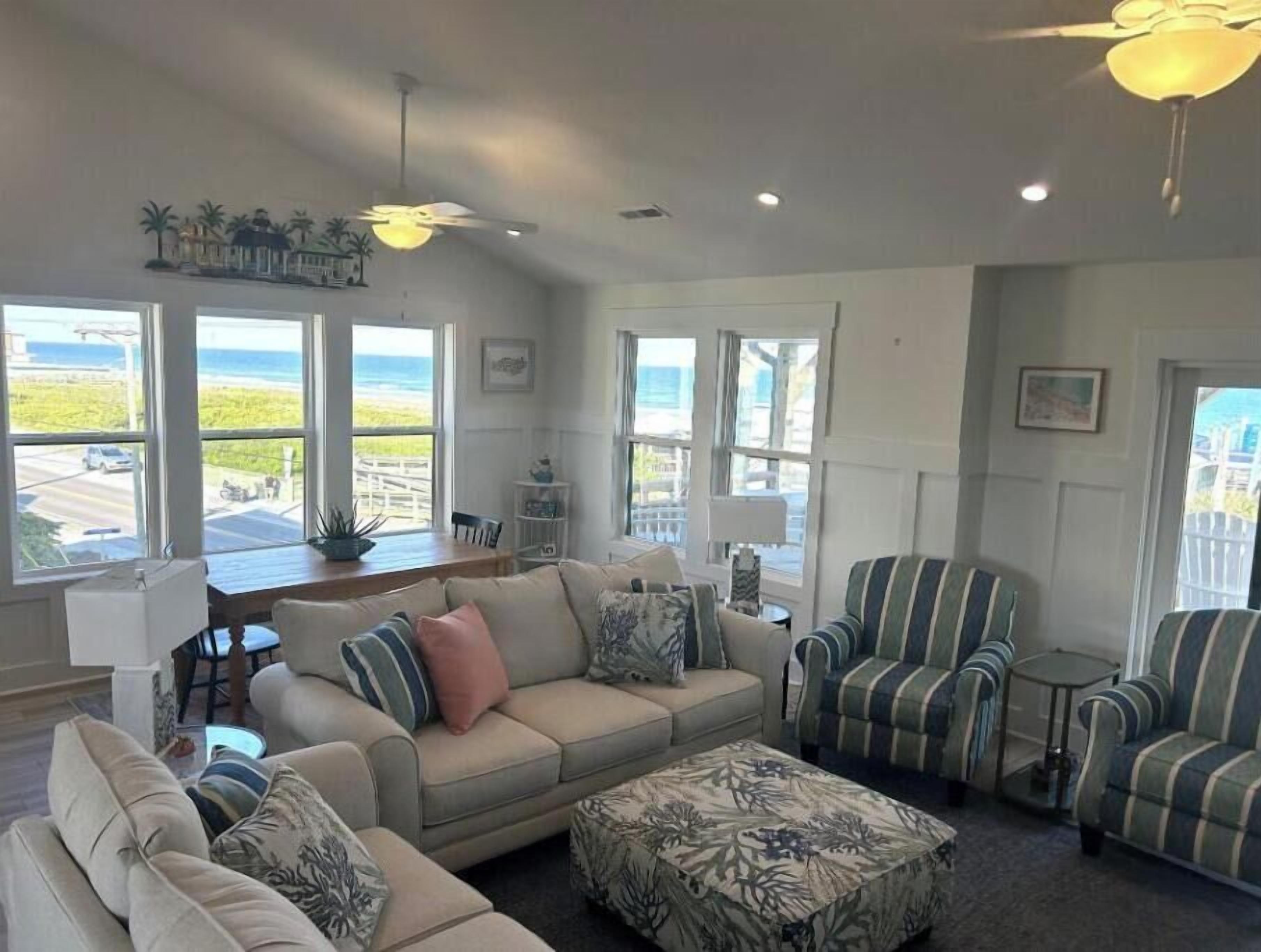 A spacious living room with a beige sofa, striped armchairs, and a patterned ottoman. Large windows offer views of the ocean and a white fence.