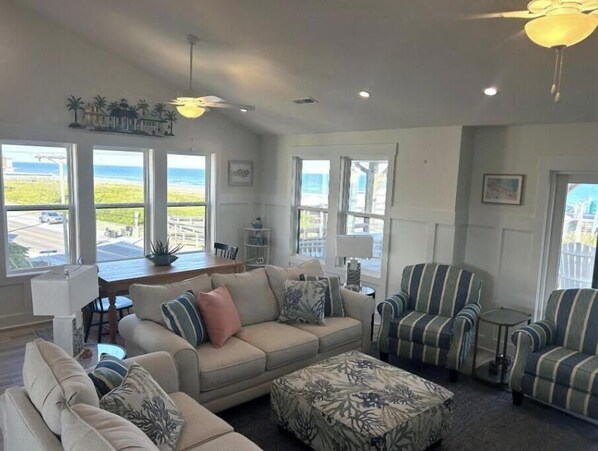 A spacious living room with a beige sofa, striped armchairs, and a patterned ottoman. Large windows offer views of the ocean and a white fence. - Bright Beach Escape • Ocean Views • Easy Beach Access (Topsail Beach,)