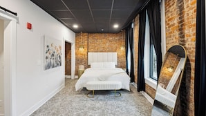Un dormitorio moderno con pared de ladrillo, cama grande, espejo y vista al exterior.