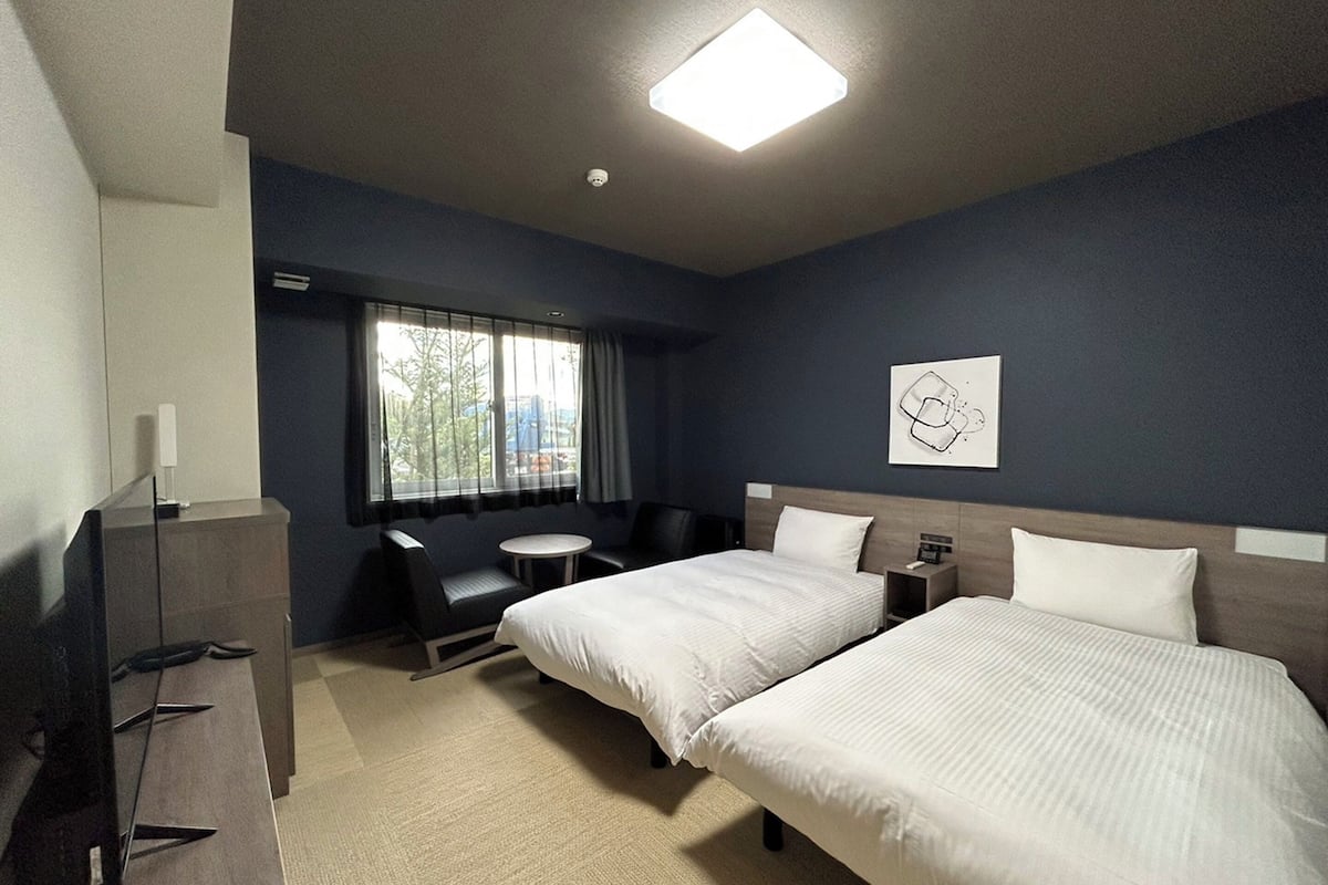 A hotel room with two beds, a desk, a chair, a small table, and a window with curtains.