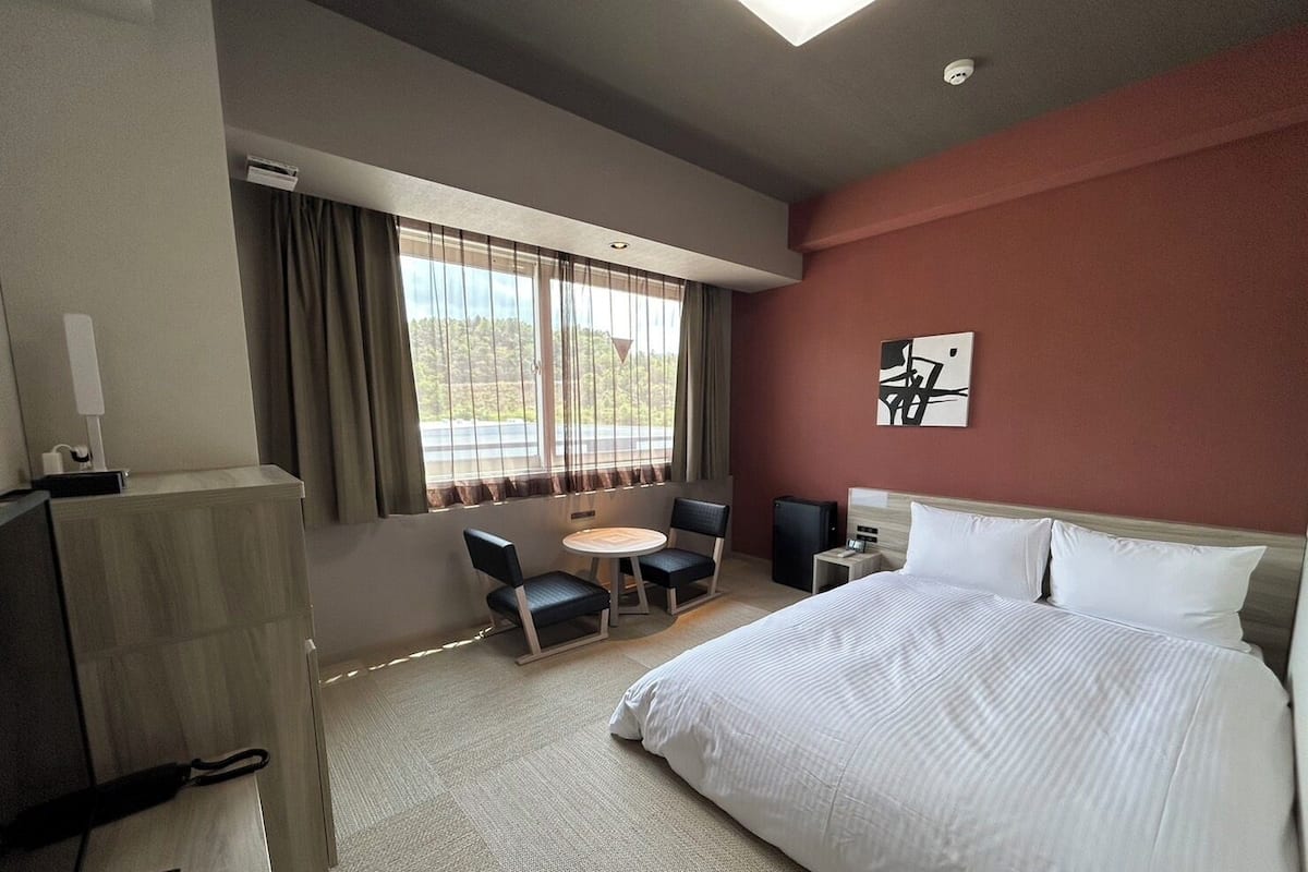 A hotel room with a bed, a desk, a chair, a small table, and a window with curtains.