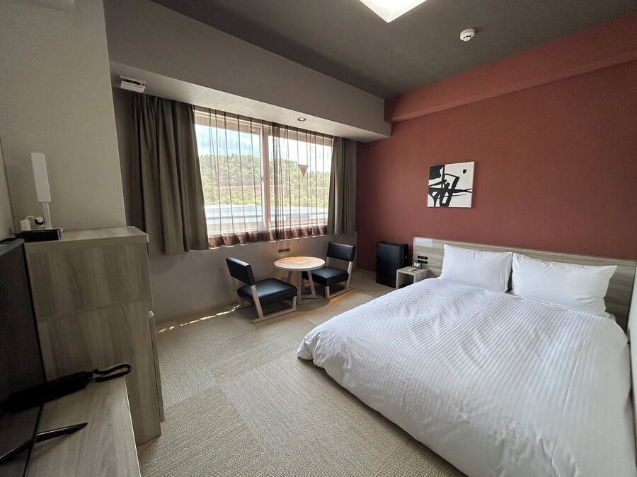 A hotel room with a bed, a desk, a chair, a small table, and a window with curtains.