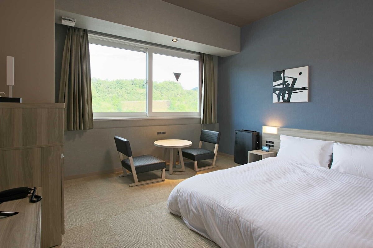 A hotel room with a bed, a desk, a chair, a small table, and a window with curtains.