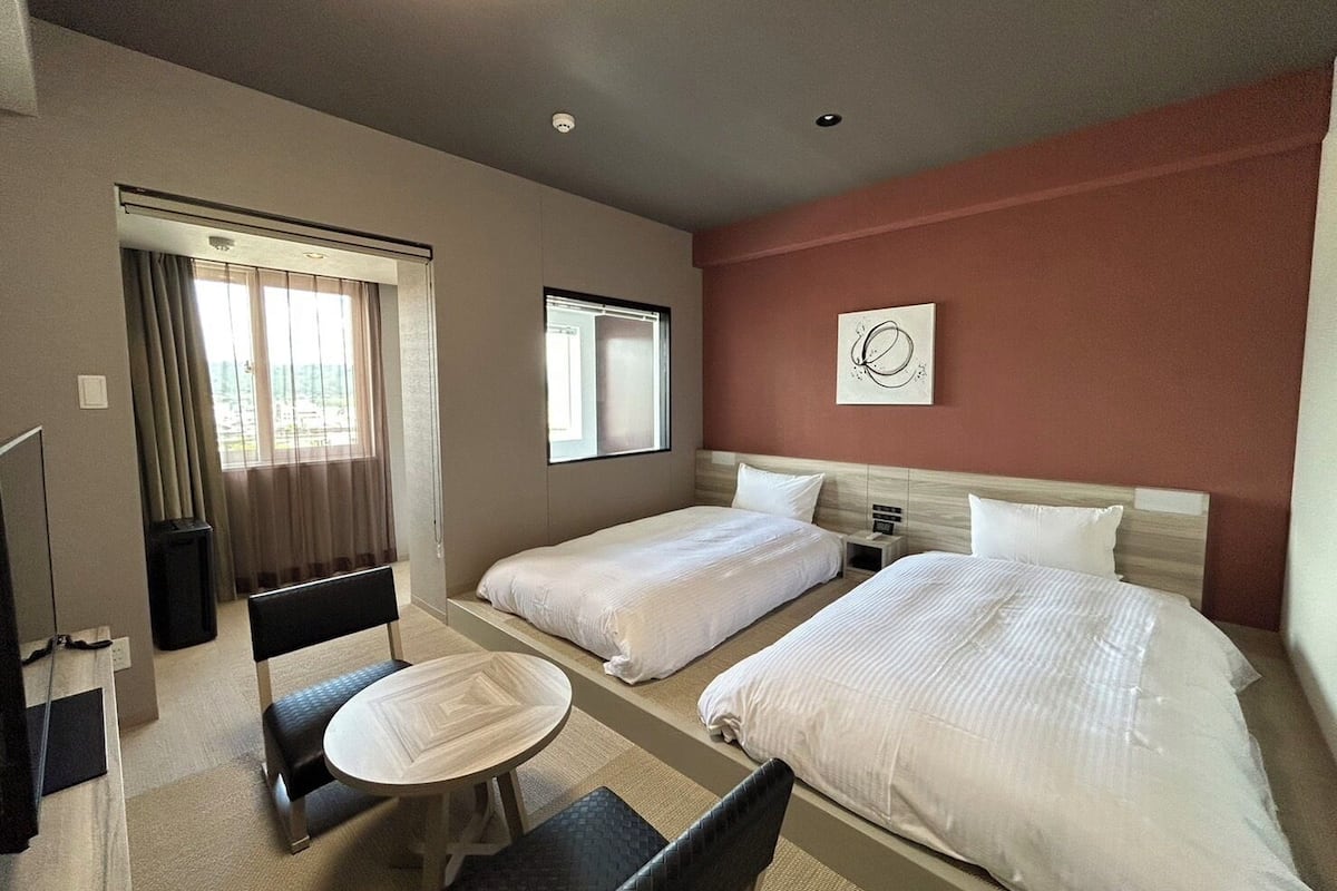 A hotel room with two beds, a small table, a chair, a TV, and a window with curtains.