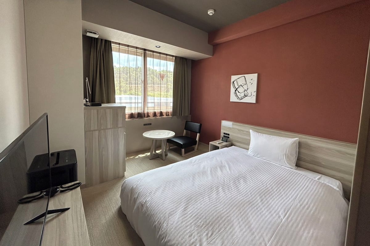 A hotel room with a bed, a desk with a computer, a chair, a small table, and a window with curtains.
