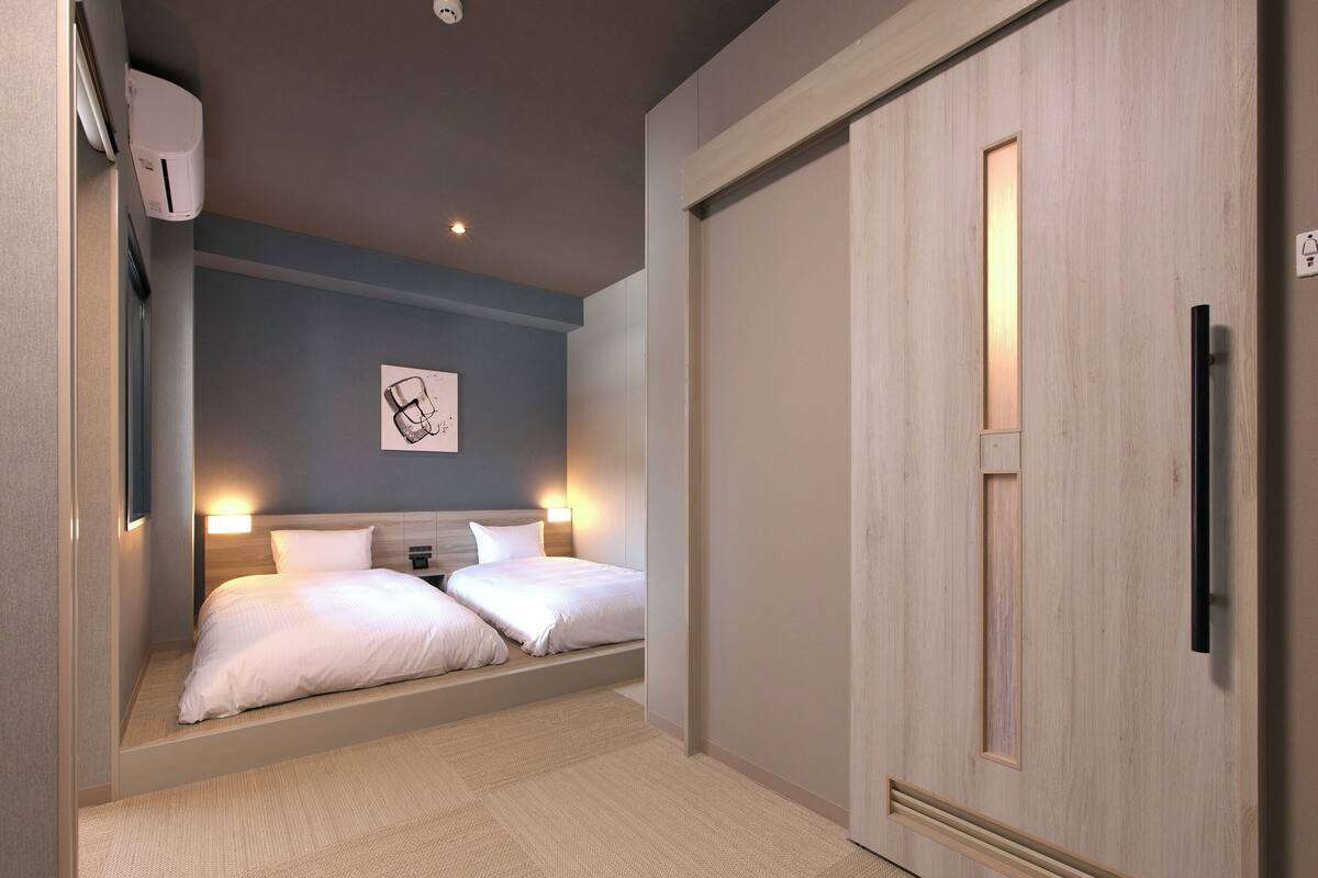 A hotel room with a bed, bedside tables, a wall-mounted air conditioner, a ceiling light, and a door with a handle.